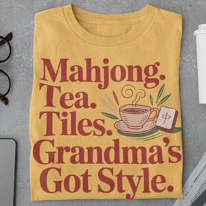 May include: Mustard yellow t-shirt with the text "Mahjong. Tea. Tiles. Grandma's Got Style." in red. The shirt features a graphic of a teacup and a Mahjong tile. Other items include glasses, a phone, headphones, and a coffee cup.
