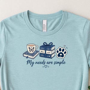 Puede incluir: Una camiseta azul claro con un estampado gráfico de una taza sobre un libro, una pila de tres libros y una huella de pata. El texto "My needs are simple" está impreso en azul oscuro debajo de las ilustraciones junto a un pequeño corazón.