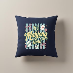 Puede incluir: Coj&iacute;n cuadrado azul marino con el texto "It's Mahjong time" en escritura amarilla. El dise&ntilde;o incluye fichas de Mahjong, dados, flores y lazos en rosa, turquesa y blanco. El fondo tiene rayas verticales turquesas.