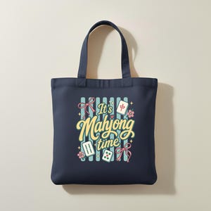 Puede incluir: Bolso tote azul marino con la frase "It's Mahjong time" en una fuente caprichosa. El dise&ntilde;o incluye fichas de Mahjong, lazos rosas y detalles florales sobre un fondo a rayas. El bolso tiene dos asas.
