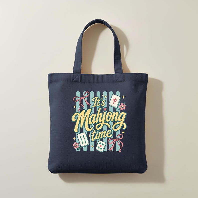 Puede incluir: Bolso tote azul marino con la frase "It's Mahjong time" en una fuente caprichosa. El dise&ntilde;o incluye fichas de Mahjong, lazos rosas y detalles florales sobre un fondo a rayas. El bolso tiene dos asas.