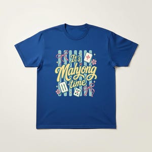 Puede incluir: Camiseta azul real con la frase "It's Mahjong Time" en escritura amarilla. El dise&ntilde;o incluye lazos rosas, flores y fichas de Mahjong. La camiseta tiene mangas cortas y cuello redondo.