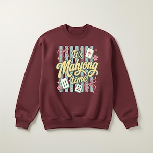 Puede incluir: Sudadera burdeos de cuello redondo con un gr&aacute;fico colorido que dice "It's Mahjong time". El dise&ntilde;o incluye fichas de Mahjong, flores y lazos. La sudadera es de color burdeos liso.