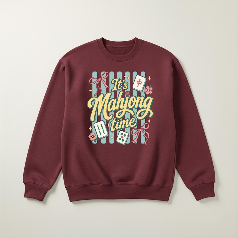 Puede incluir: Sudadera burdeos de cuello redondo con un gr&aacute;fico colorido que dice "It's Mahjong time". El dise&ntilde;o incluye fichas de Mahjong, flores y lazos. La sudadera es de color burdeos liso.