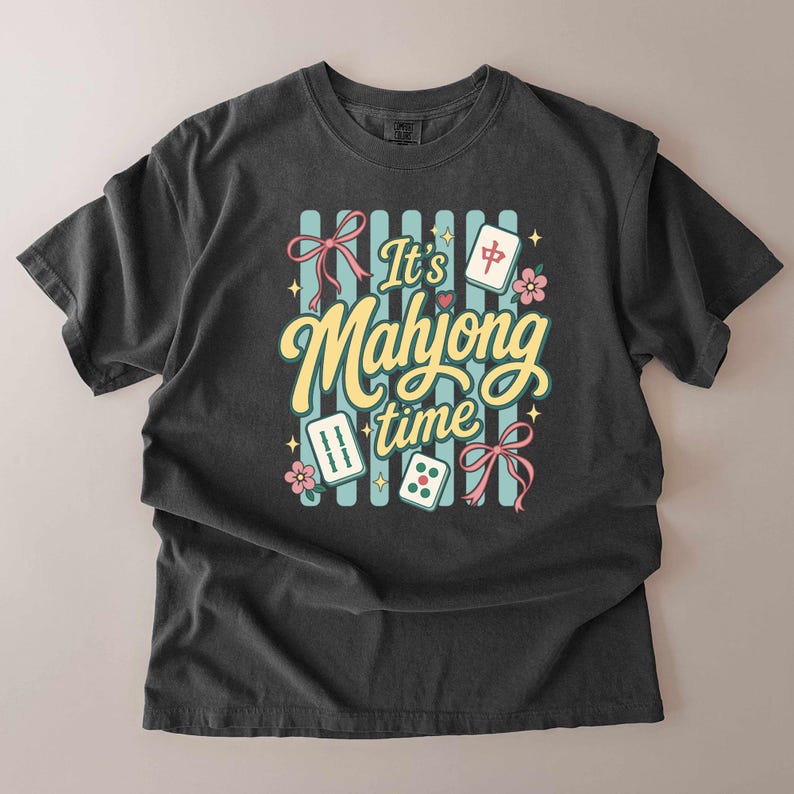 Puede incluir: Camiseta gris oscuro con la frase "It's Mahjong time" en un estilo retro. El dise&ntilde;o incluye fichas de mahjong, dados, flores y lazos sobre un fondo a rayas. La camiseta est&aacute; hecha de un material suave y c&oacute;modo.