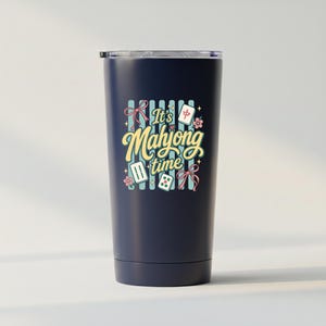 Puede incluir: Un vaso t&eacute;rmico azul marino con tapa. El vaso presenta el texto "It's Mahjong time" en una fuente caprichosa, junto con ilustraciones de fichas de mahjong, lazos y flores. El dise&ntilde;o est&aacute; sobre un fondo a rayas.