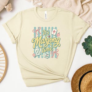 Puede incluir: Camiseta color crema con la frase "It's Mahjong Time" en una fuente retro, con ilustraciones de fichas de Mahjong, dados y lazos. El dise&ntilde;o incluye un fondo a rayas azules y detalles florales, creando una est&eacute;tica divertida.