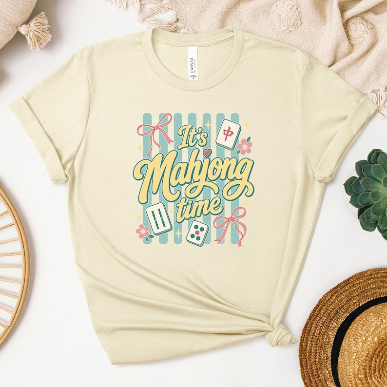 Puede incluir: Camiseta color crema con la frase "It's Mahjong Time" en una fuente retro, con ilustraciones de fichas de Mahjong, dados y lazos. El dise&ntilde;o incluye un fondo a rayas azules y detalles florales, creando una est&eacute;tica divertida.