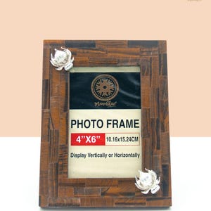 May include: A brown wooden photo frame with a 4"x6" opening. The frame has a decorative design and is decorated with silver metal flowers. The text "PHOTO FRAME 4"x6" 10.16x15.24CM Display Vertically or Horizontally" is printed on the frame.