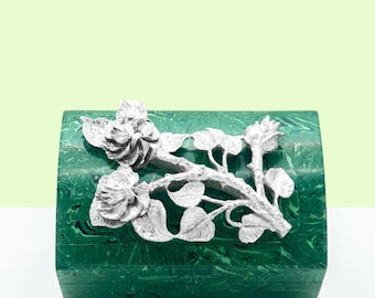 May include: A green malachite box with a silver rose design on the lid. The box is closed and sitting on a white surface.