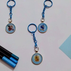 May include: Three blue keychains with cartoon character images. Each keychain has a round, light blue pendant with a different character: Goofy and Mickey Mouse, Donald Duck, and Pluto. The keychains are on a white surface with blue markers.
