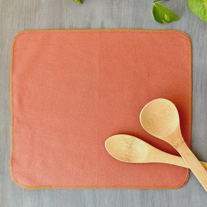 May include: A rectangular, coral-colored dish drying mat with a stitched, golden-yellow border. Two wooden spoons rest on the mat. The mat is made of a soft, absorbent material, perfect for kitchen use.