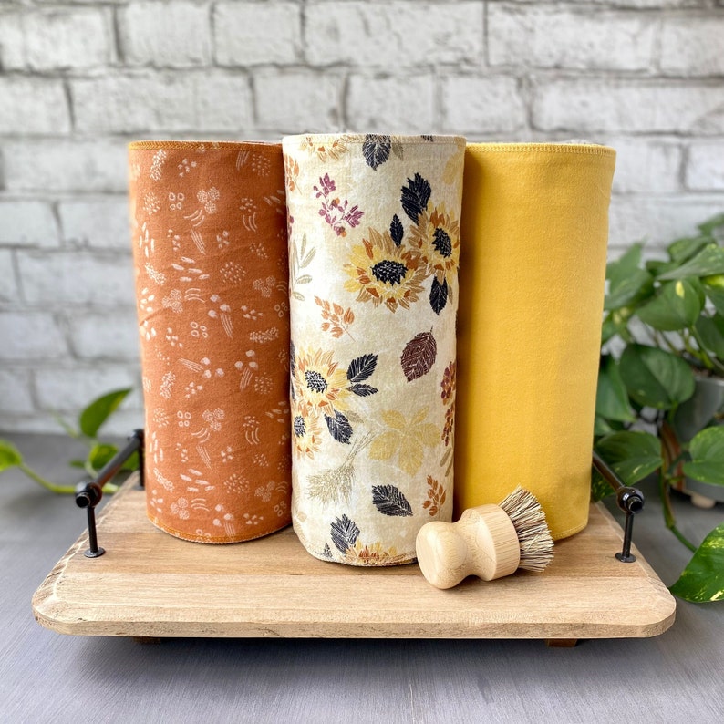 May include: Three rolled-up kitchen towels with autumn-themed patterns and a wooden dish brush on a wooden tray. The towels feature floral and sunflower designs in shades of orange, cream, and yellow. The dish brush has a wooden handle and bristles.