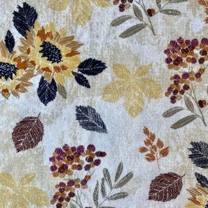 May include: A cream-colored fabric with a repeating pattern of sunflowers, leaves, and floral elements in shades of yellow, brown, and burgundy. The design includes detailed leaf and flower illustrations.