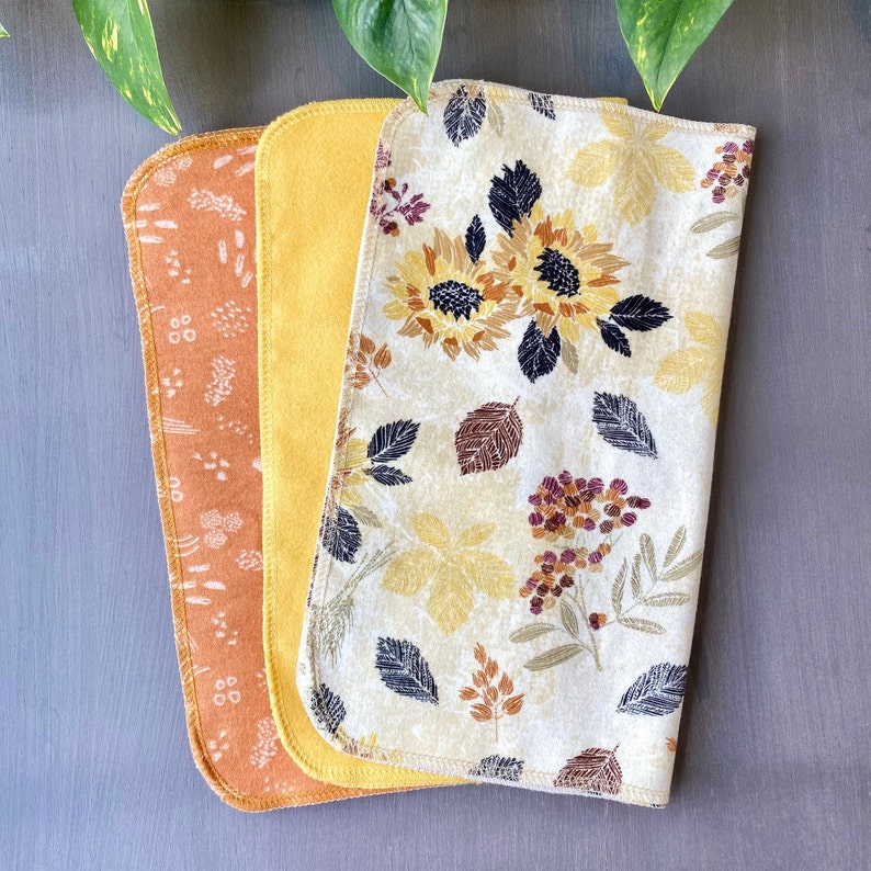 May include: Three rectangular cloth items. One is orange with a white floral pattern, the second is solid yellow, and the third is cream-colored with a sunflower and leaf design. The items are stacked on a gray surface.