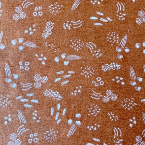 May include: Close-up of a textured, rust-colored fabric with a repeating pattern of white abstract shapes. The design includes small dots, curved lines, and clusters of dots, creating a visually interesting surface.