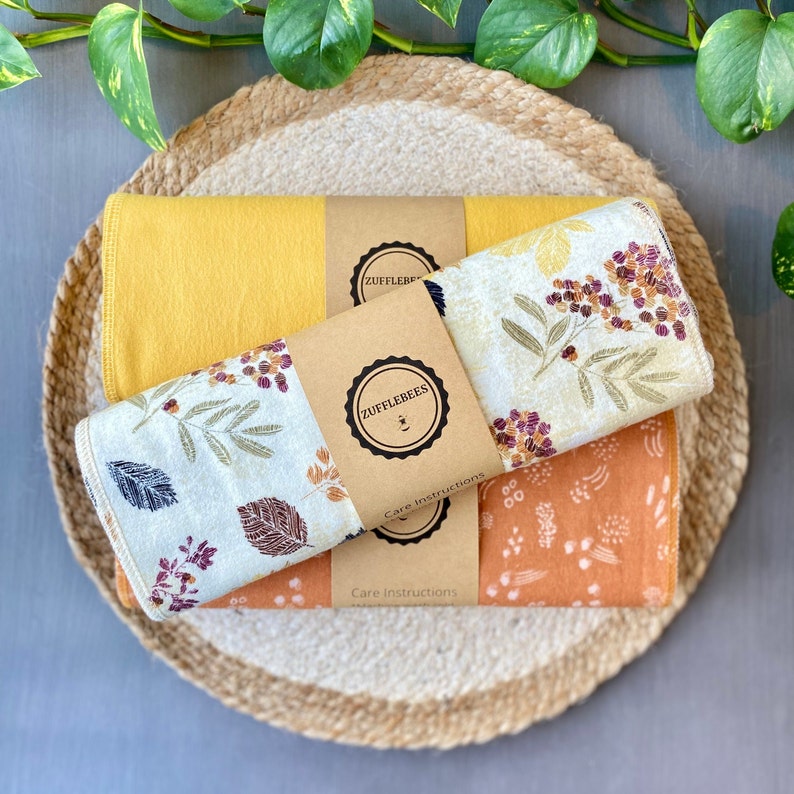 May include: A stack of three cloth napkins in shades of yellow, cream, and orange, with botanical patterns. The napkins are wrapped with brown paper bands featuring the "ZUFFLEBEES" logo and "Care Instructions" text. The napkins are on a woven placemat.