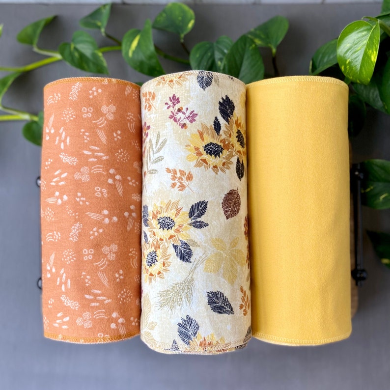 May include: Three rolled fabric pieces. One is orange with a white floral pattern, the second is cream with sunflowers and leaves, and the third is solid yellow. The fabrics are displayed vertically.