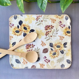 May include: A rectangular beige kitchen mat with a floral pattern featuring sunflowers, leaves, and berries in yellow, brown, and purple. Two wooden spoons rest on the mat, which is placed on a gray surface.