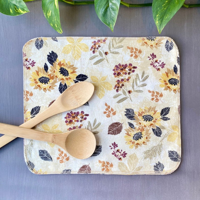 May include: A rectangular beige kitchen mat with a floral pattern featuring sunflowers, leaves, and berries in yellow, brown, and purple. Two wooden spoons rest on the mat, which is placed on a gray surface.