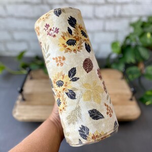 May include: A cylindrical lampshade featuring a cream-colored background with a floral pattern. The design includes sunflowers, leaves, and wheat stalks in shades of yellow, brown, and black. The lampshade is a home decor item.