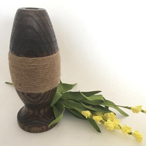 May include: A dark brown wooden vase with a natural twine band around the middle. The vase is oval shaped and has a wide base. The vase is sitting on a white surface with a bouquet of yellow flowers.