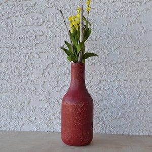 May include: A red glass bottle vase with a textured surface, holding a bouquet of yellow flowers and green leaves.