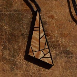 Peut inclure: Collier pendentif géométrique en bois. Le pendentif est de forme triangulaire avec un bord noir et un intérieur marron clair à motifs. Le cordon du collier est noir.