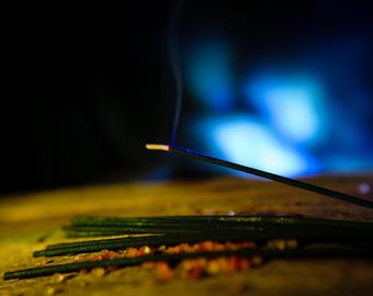 May include: Close-up of incense sticks with smoke rising against a blurred blue and black background. Several green incense sticks are on a wooden surface with small orange granules. The lit incense stick has a glowing tip.