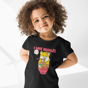 May include: A black t-shirt featuring a cartoon illustration of a yellow cat enjoying a bowl of noodles. The text "I LOVE NOODLES" is printed in pink above the cat. The cat has a speech bubble with the text "食 物" in pink.