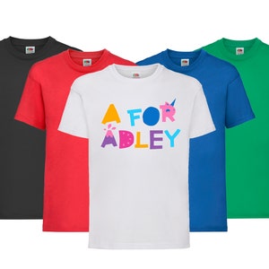 K&ouml;nnte beinhalten: F&uuml;nf T-Shirts in verschiedenen Farben: Schwarz, Rot, Wei&szlig;, Blau und Gr&uuml;n. Das wei&szlig;e T-Shirt hat einen bunten Aufdruck mit der Aufschrift "A for Adley".