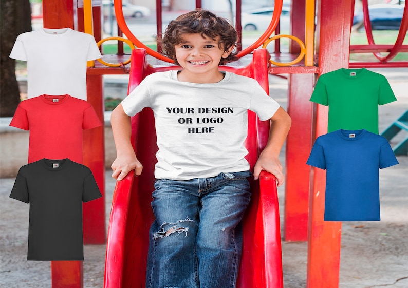 K&ouml;nnte beinhalten: Ein junges Kind tr&auml;gt ein wei&szlig;es T-Shirt mit dem Aufdruck "YOUR DESIGN OR LOGO HERE". Das Kind sitzt auf einer roten Rutsche. Im Hintergrund sind f&uuml;nf leere T-Shirts in verschiedenen Farben zu sehen.