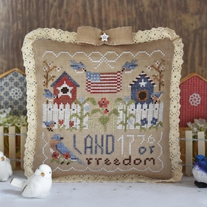 May include: A square, beige, cross-stitched pillow with a patriotic theme. It features a stitched American flag, birdhouses, birds, and the words "LAND of freedom 1776". The pillow is trimmed with lace and a burlap bow.