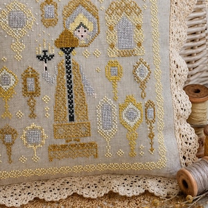 Puede incluir: Una tela de lino beige con un patr&oacute;n de punto de cruz de una figura central que sostiene un candelabro, rodeada de varios dise&ntilde;os de espejos ornamentados en hilo dorado y plateado. La tela est&aacute; rematada con un borde de encaje color crema.