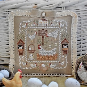 May include: A square, beige cross-stitch pillow with a farm scene. The design features chickens, a farmhouse, and the text "Farm Fresh Eggs". The pillow has a lace trim and a decorative bow at the top.