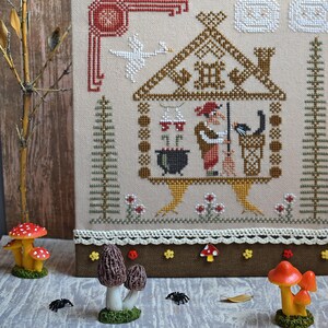 May include: Cross-stitch embroidery featuring a witch in a house with a black cat, cauldron, and goose. The embroidery is framed with a brown border and decorated with mushroom and spider figurines. The color palette includes beige, brown, red, and black.