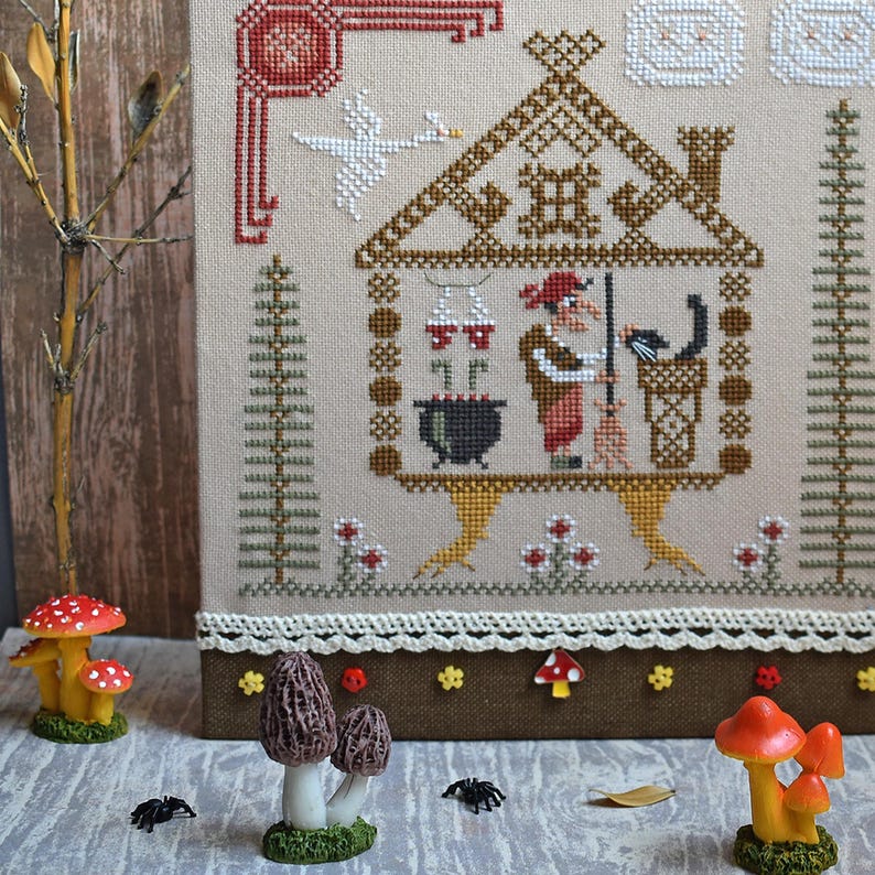 May include: Cross-stitch embroidery featuring a witch in a house with a black cat, cauldron, and goose. The embroidery is framed with a brown border and decorated with mushroom and spider figurines. The color palette includes beige, brown, red, and black.
