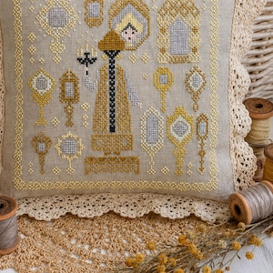 Puede incluir: Un coj&iacute;n cuadrado de lino con un dise&ntilde;o de punto de cruz que muestra una figura central sosteniendo un candelabro, rodeada de varios espejos de mano ornamentados en hilos dorados y plateados. El coj&iacute;n tiene un borde de encaje color crema.