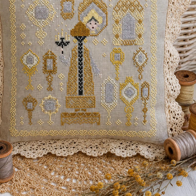 Puede incluir: Un coj&iacute;n cuadrado de lino con un dise&ntilde;o de punto de cruz que muestra una figura central sosteniendo un candelabro, rodeada de varios espejos de mano ornamentados en hilos dorados y plateados. El coj&iacute;n tiene un borde de encaje color crema.