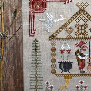 May include: Cross-stitch sampler featuring a whimsical scene with a house, goose, and witch. The design uses brown, red, green, and white thread on a beige background. A tree and flowers are also included in the design.