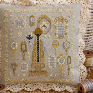 Puede incluir: Un coj&iacute;n cuadrado de color beige con un dise&ntilde;o de punto de cruz que muestra una figura central sosteniendo un candelabro, rodeada de varios espejos de mano y marcos ornamentados en hilo dorado, plateado y negro, con un borde de encaje de ganchillo.