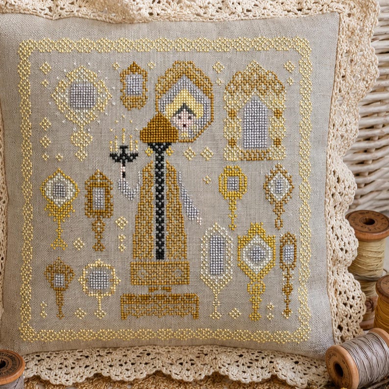Puede incluir: Un coj&iacute;n cuadrado de color beige con un dise&ntilde;o de punto de cruz que muestra una figura central sosteniendo un candelabro, rodeada de varios espejos de mano y marcos ornamentados en hilo dorado, plateado y negro, con un borde de encaje de ganchillo.