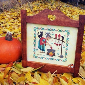 May include: A framed cross-stitch design featuring a witch stirring a cauldron with a black cat and a raven in the background. The witch is wearing a red and blue dress and a white hat. The background is a cream color with a green border.