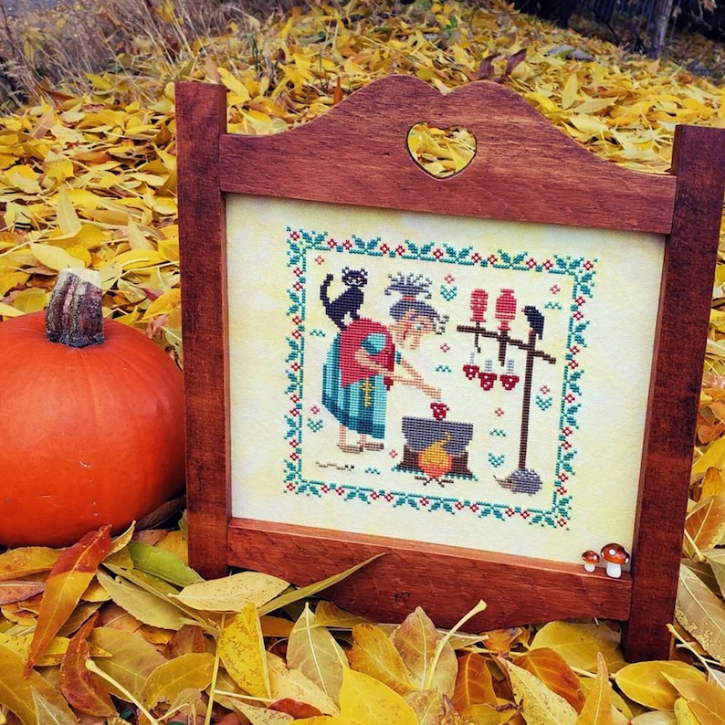 May include: A framed cross-stitch design featuring a witch stirring a cauldron with a black cat and a raven in the background. The witch is wearing a red and blue dress and a white hat. The background is a cream color with a green border.