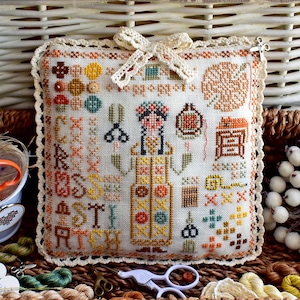 May include: A white pillow with a cross-stitch design featuring a woman in a floral dress, scissors, and other needlework tools. The pillow has a crocheted border and a brown bow.
