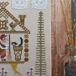 May include: Cross-stitch embroidery featuring a whimsical scene with a house, a witch, and a black cat. The design uses brown, red, green, and beige threads. The artwork is framed by a green border and small red and white flowers.