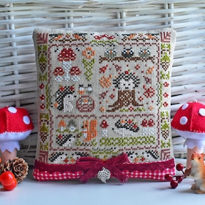 May include: A square-shaped pillow with a cross-stitch design featuring a woodland scene with mushrooms, birds, squirrels, and hedgehogs. The pillow has a red and white checkered fabric border and a red bow with a silver pin.