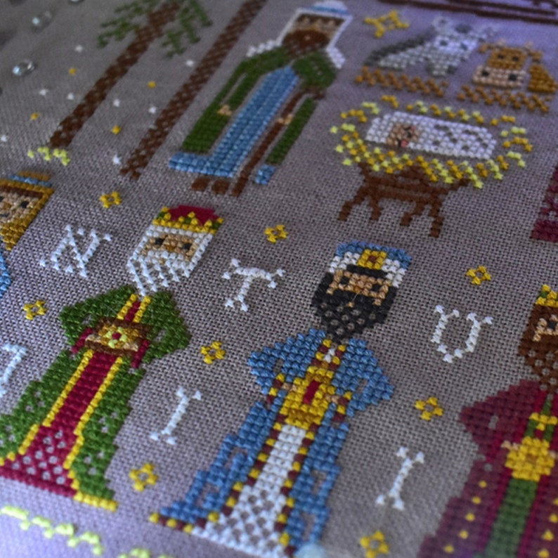 K&ouml;nnte beinhalten: Eine detaillierte Kreuzsticharbeit, die eine Weihnachtskrippe darstellt. Das Design zeigt Figuren der Heiligen Drei K&ouml;nige, das Jesuskind in einer Krippe und andere biblische Figuren. Die Farbpalette umfasst Schattierungen von Gr&uuml;n, Blau, Rot und Gold auf einem neutralen Hintergrund.