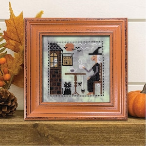 May include: A square cross-stitch embroidery in a distressed orange wooden frame. The design features a witch in a black dress and hat sitting at a table with a black cat, a sign reading Salem Brew, a moon, and two spiders on a light green background.