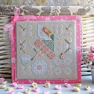 May include: A cross-stitch embroidery design featuring a fairy with pink and green wings, sitting on a flower. The design is surrounded by flowers and butterflies, and is framed in a pink border.